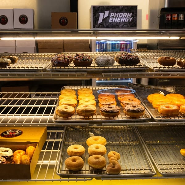 Display case filled with assorted freshly baked bagels, donuts, and pastries on trays, with a Phorm Energy sign behind.