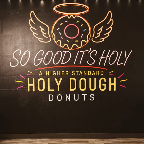 Neon-style Holy Dough Donuts mural with a winged halo donut, surrounded by dark restaurant seating.