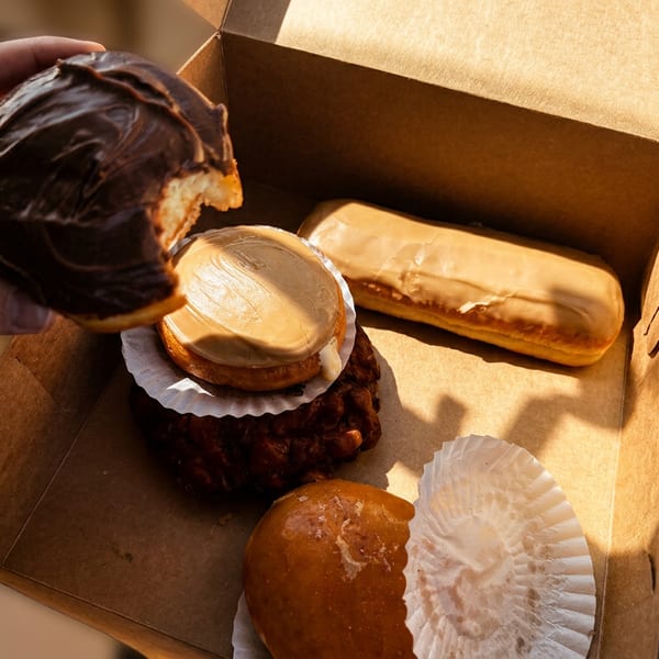 A hand holds a chocolate-glazed pastry over an open box containing assorted glazed doughnuts and pastries.
