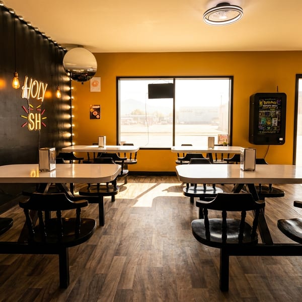 Warmly lit restaurant dining area with empty tables, black chairs, wood flooring, pendant lights, and a neon Holy Sh sign.