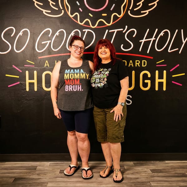 Two smiling women pose beneath a colorful “So Good It’s Holy” mural inside a warmly lit restaurant.
