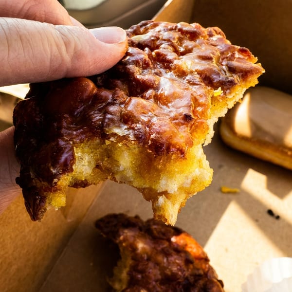 Hand holding a partially eaten pecan pastry with glossy caramelized topping over a box of additional pastries.